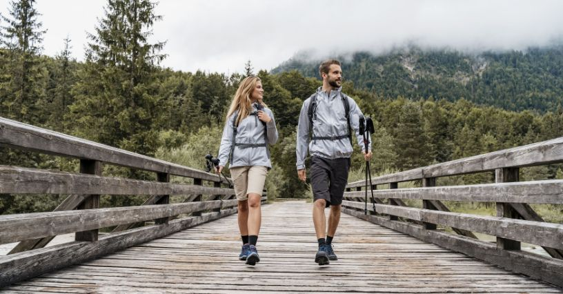 Două persoane merg pe un pod din lemn, îmbrăcate în echipament de drumeție