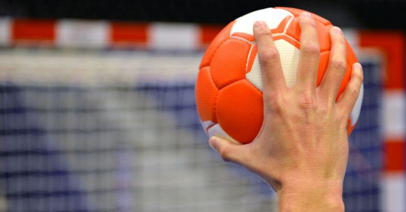 Mâna unei persoane ține o minge de handbal în fața unei porți