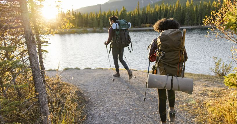 Două persoane fac drumeții lângă un lac, purtând echipament outdoor