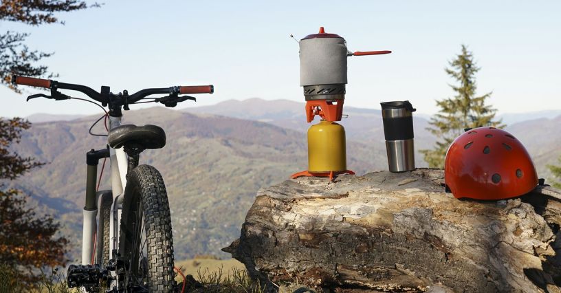 O bicicletă și un aragaz turistic sunt așezate pe un trunchi în fața unui peisaj montan